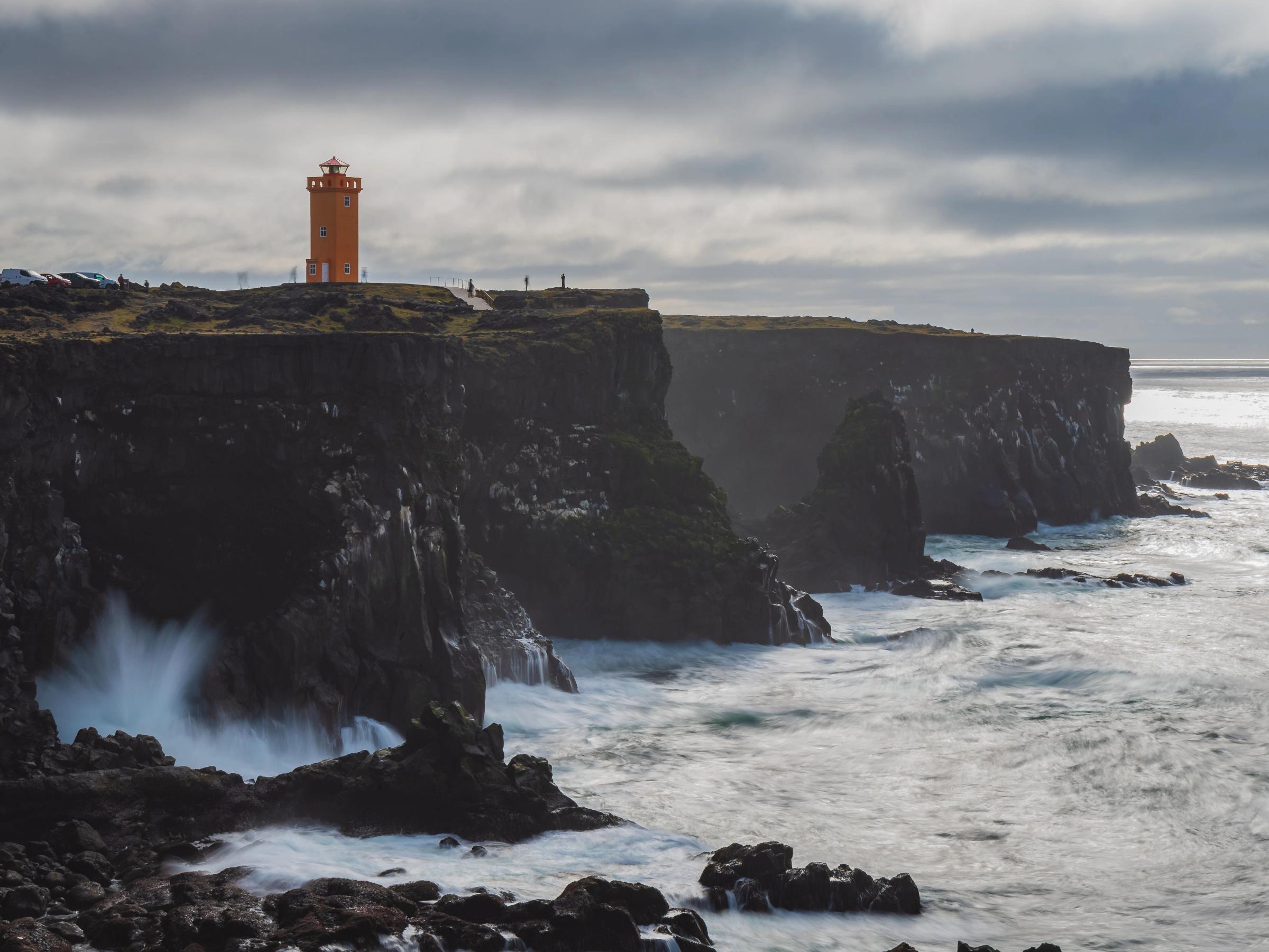 paisaje con acantilados donde rompen las olas del mar con un faro naranja encima
