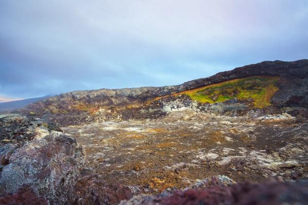 A volcanic crater with a dark rocky floor, vibrant green and yellow growth on the far wall, under a cloudy sky.