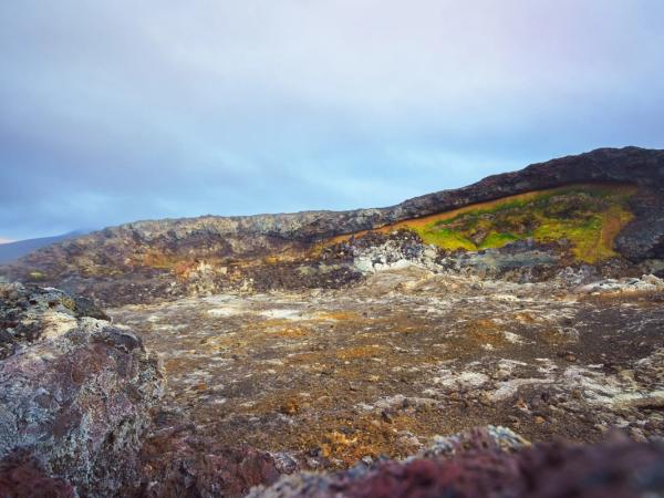 A geothermal landscape with dark, rocky ground, pale mineral deposits, and a vibrant green moss patch on a distant rock wall.