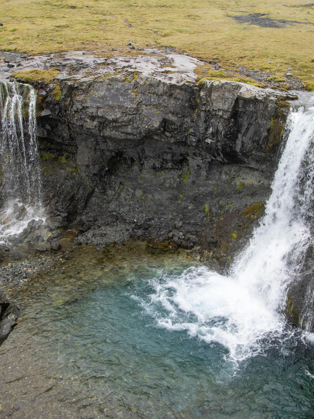 Two waterfalls cascade into a clear blue pool surrounded by rocky cliffs.