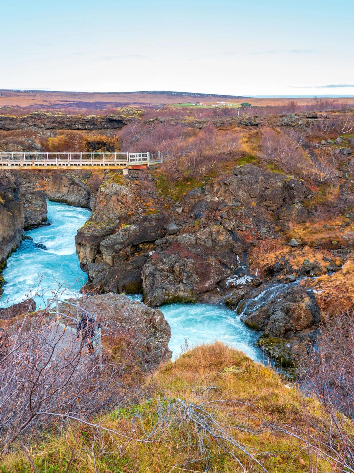 A vibrant blue river flows through a rocky canyon, spanned by a footbridge, surrounded by sparse reddish-brown autumn vegetation.