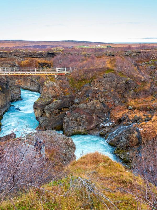 A vibrant blue river flows through a rocky canyon, spanned by a footbridge, surrounded by sparse reddish-brown autumn vegetation.