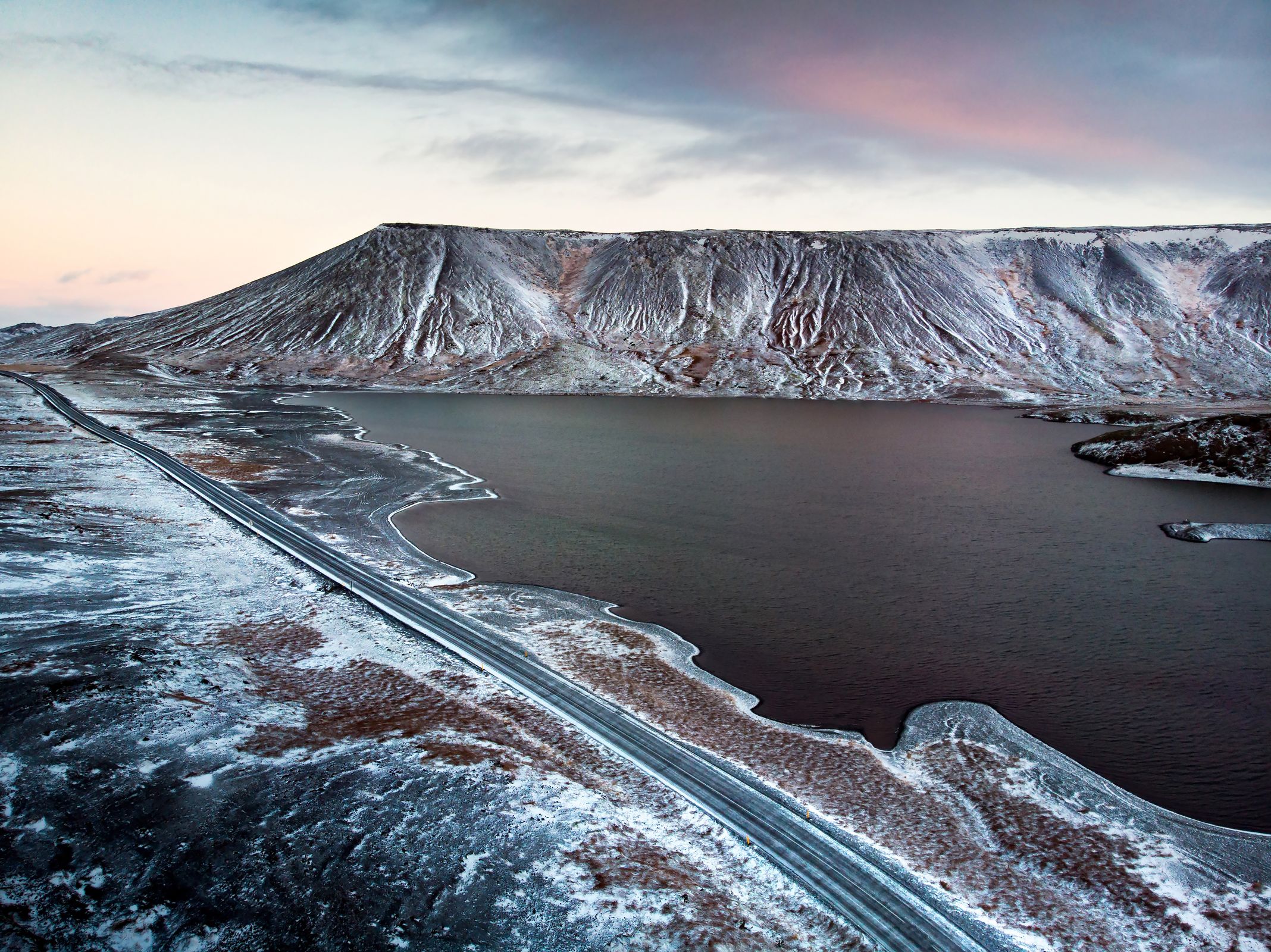 an aerial view of a road leading to a lake surrounded by snow covered mountains in iceland.