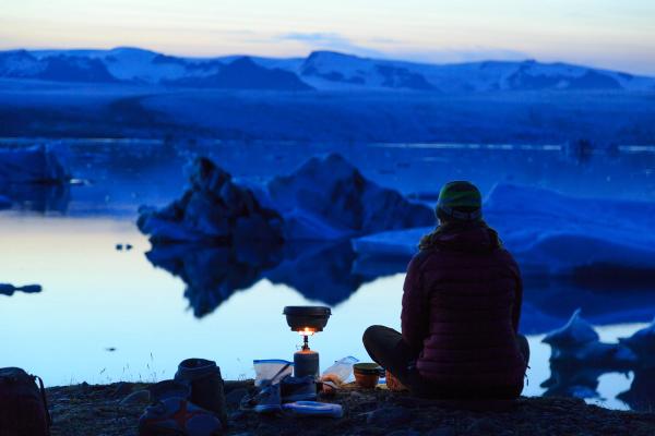 A person cooks on a portable stove beside a glacial lake with icebergs and mountains in the distance at dusk.