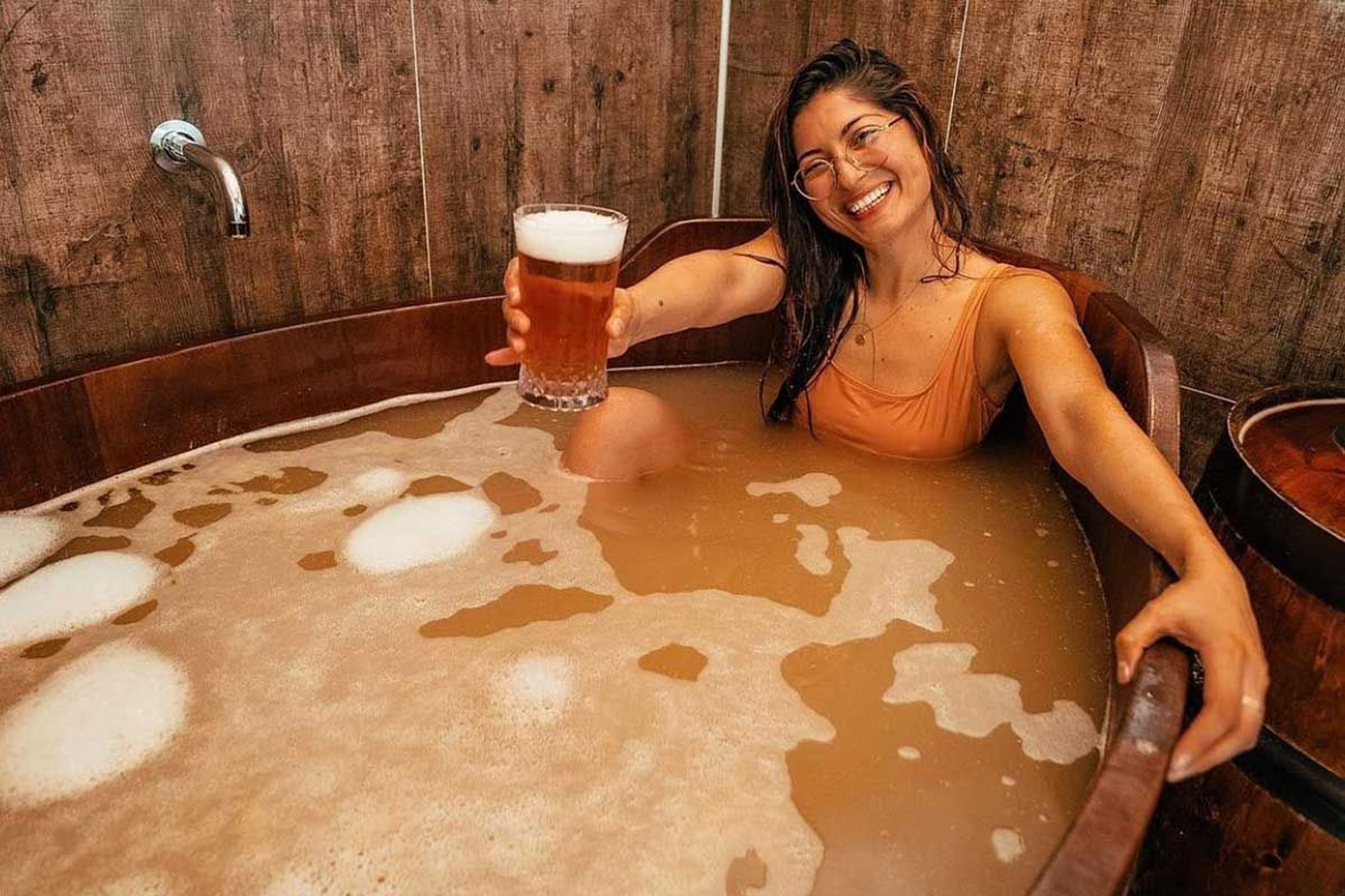 Smiling woman in a wooden tub filled with foamy beer, holding up a glass of beer.