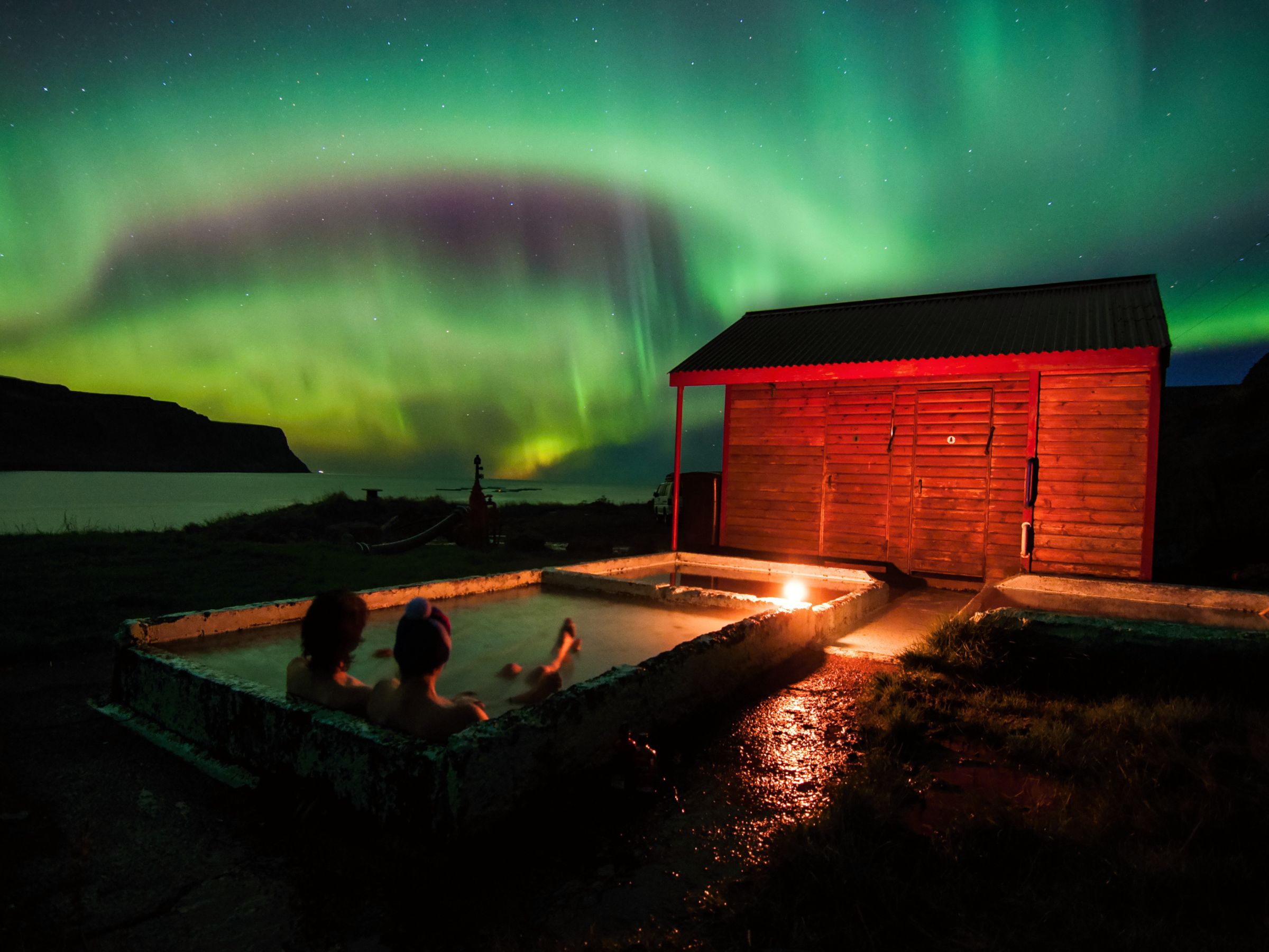 People relaxing in a small geothermal pool under the northern lights in Iceland, beside a red wooden cabin.