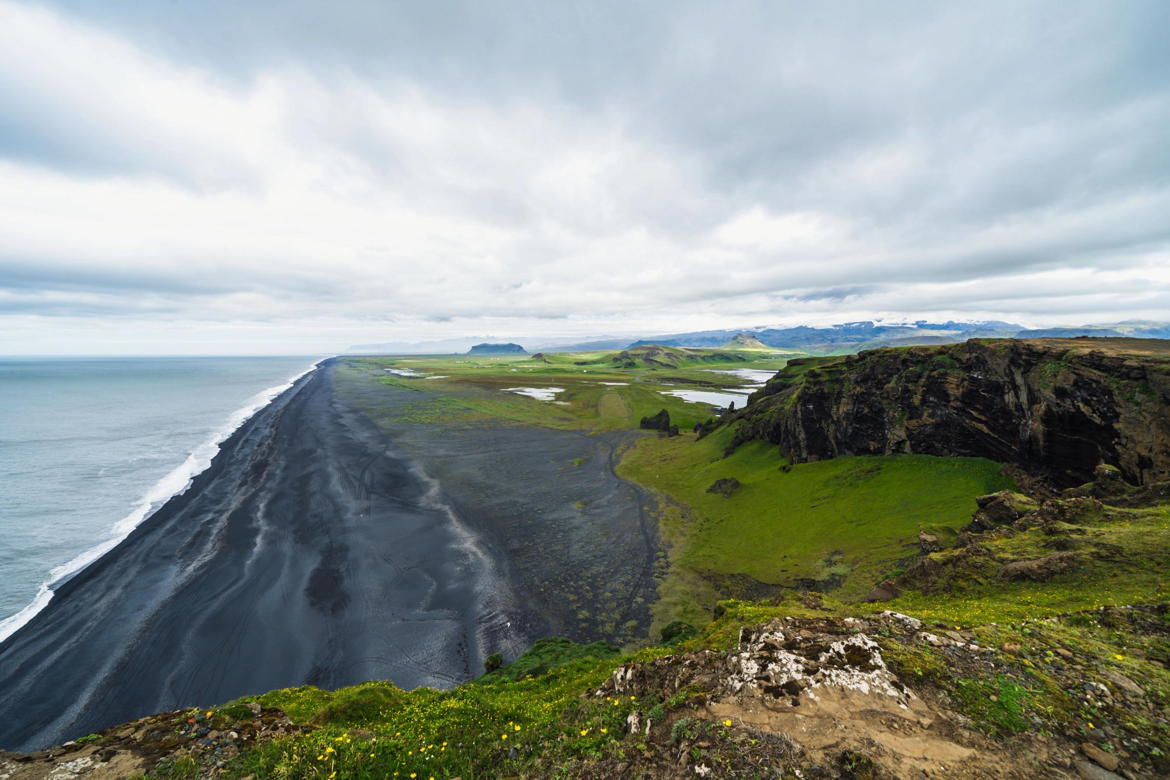 A long black sand beach meets the ocean, flanked by green cliffs and plains under a cloudy sky.