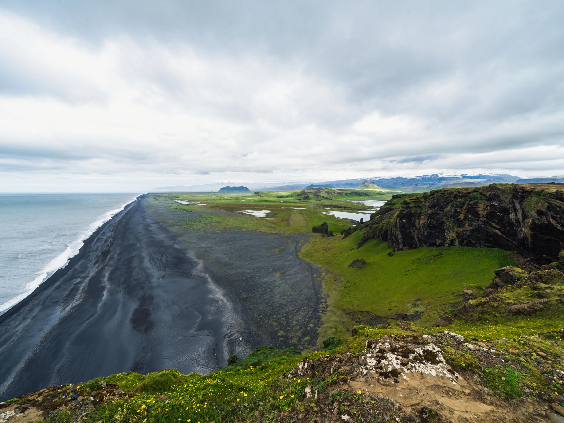 A long black sand beach meets the ocean, flanked by green cliffs and plains under a cloudy sky.