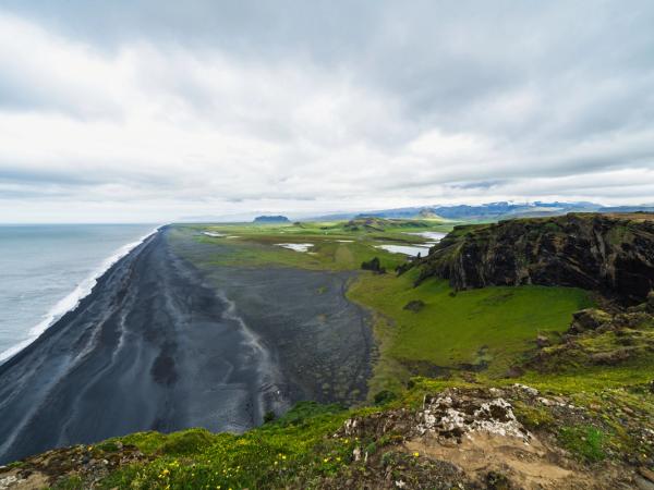 Una larga playa de arena negra se encuentra con el océano, flanqueada por acantilados verdes y llanuras bajo un cielo nublado.
