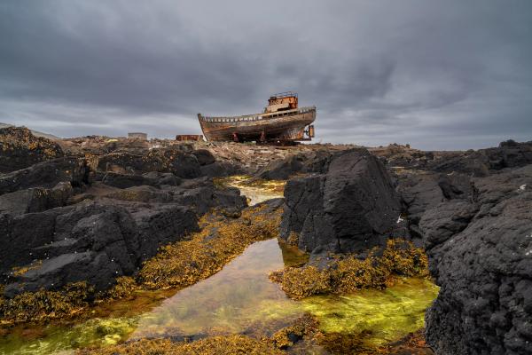 Rusty shipwreck on a dark, rocky shore with tide pools under a cloudy sky.