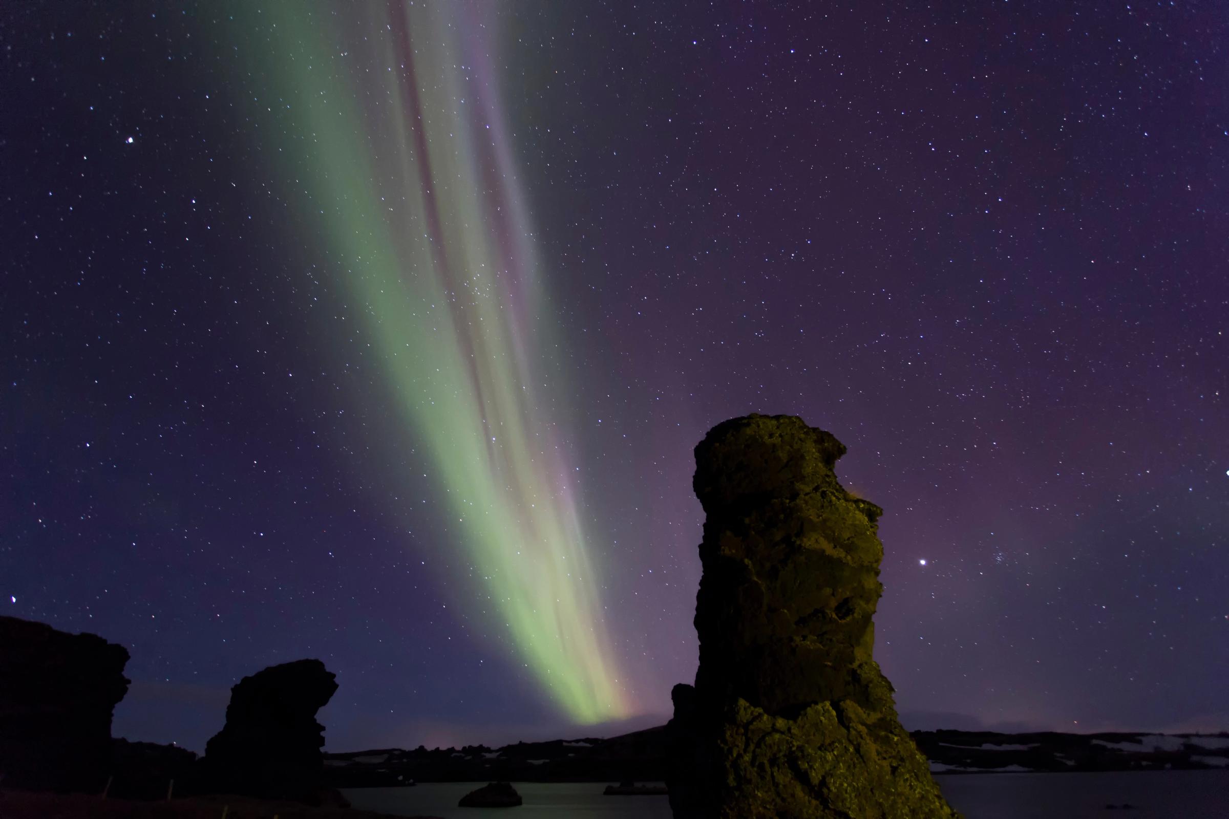 Cortinas de aurora boreal verde y morada se extienden por un cielo oscuro y estrellado sobre formaciones rocosas en silueta.