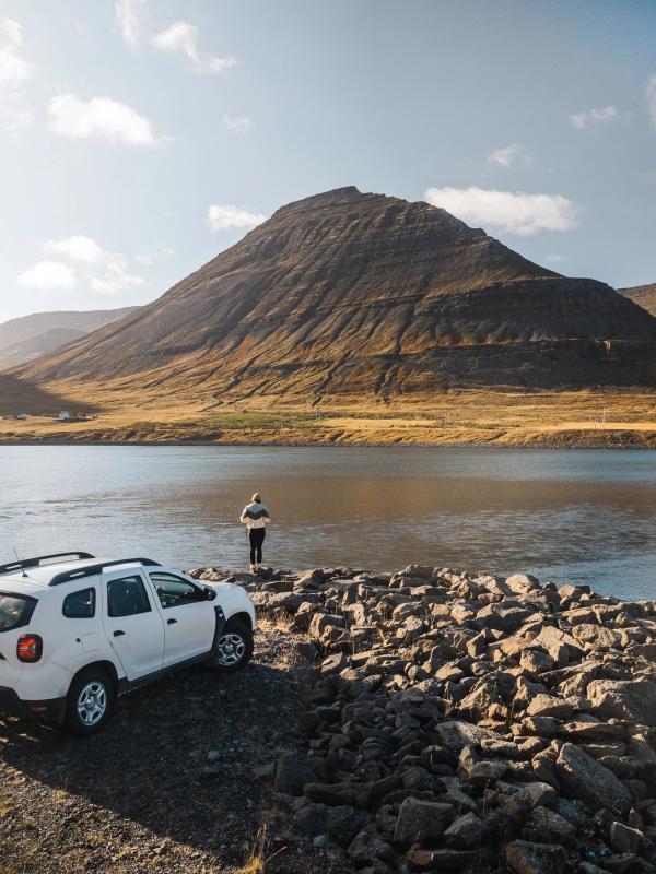 A white SUV parked on a rocky shore next to a person looking across a lake at a large, barren mountain under a bright sky.