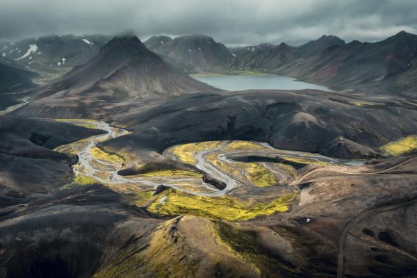 Aerial view of dark volcanic mountains with a winding river bordered by bright green moss and a distant lake under a cloudy sky.