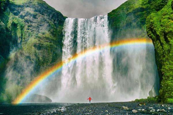 a person in a red jacket standing in front of a waterfall with a rainbow
