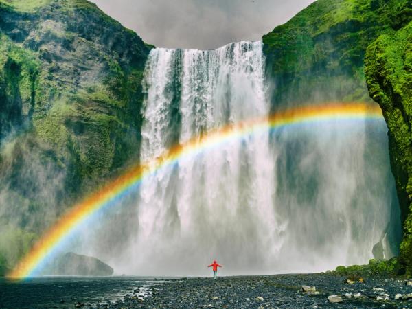 Une personne se tient devant une cascade avec un arc-en-ciel en arrière-plan.