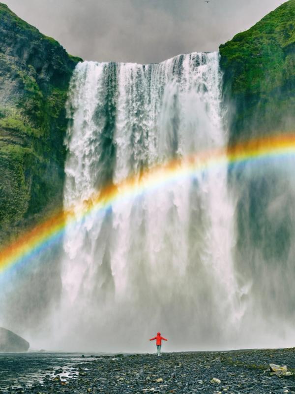 A vibrant rainbow arcs in front of a powerful waterfall cascading down lush green cliffs, with a small person in red standing at the base.