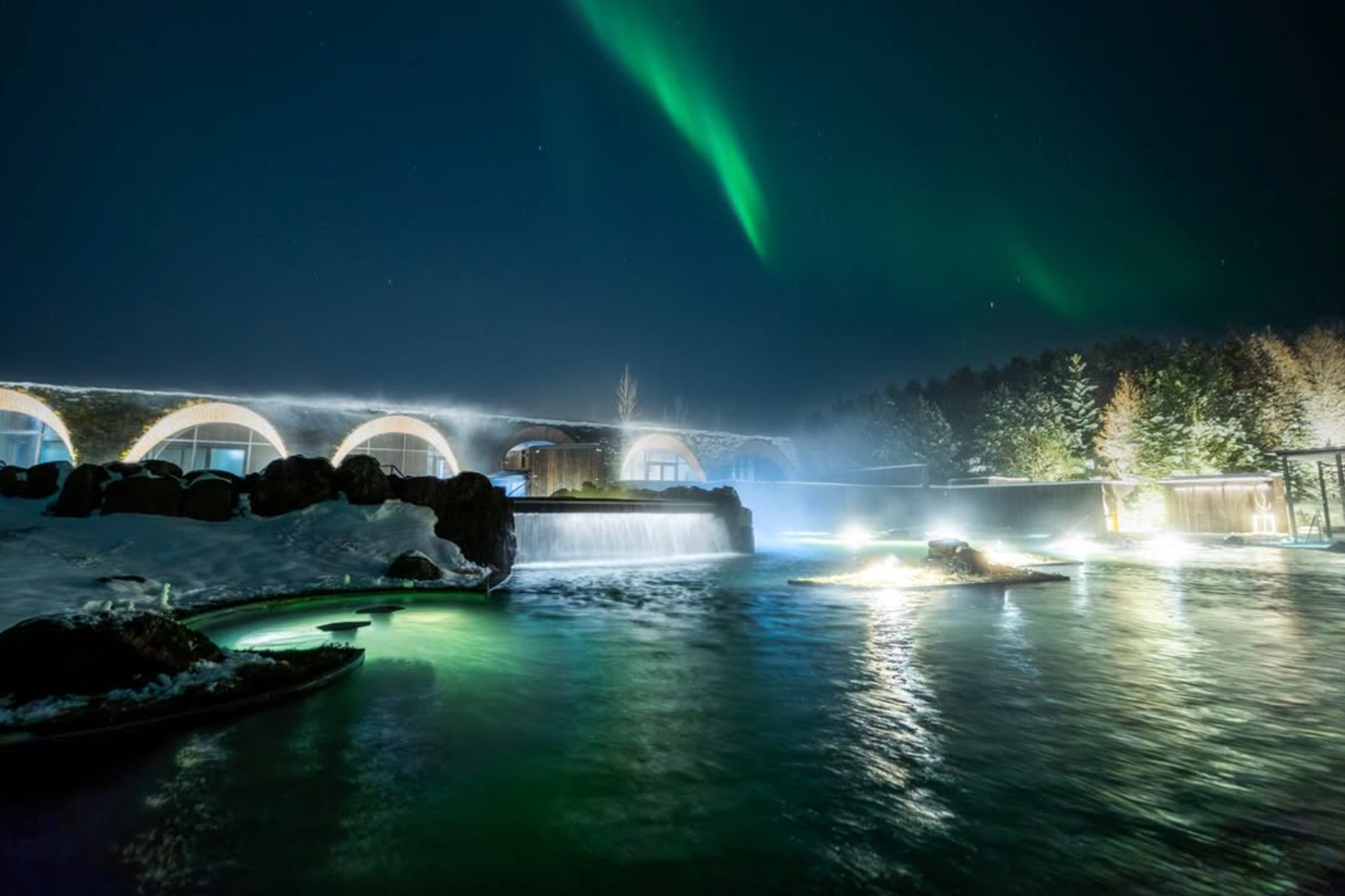 Las auroras boreales brillan sobre una humeante laguna termal al aire libre y un edificio moderno en la noche.