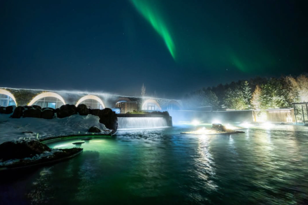 Northern Lights glow above a steamy outdoor thermal lagoon and modern building at night.