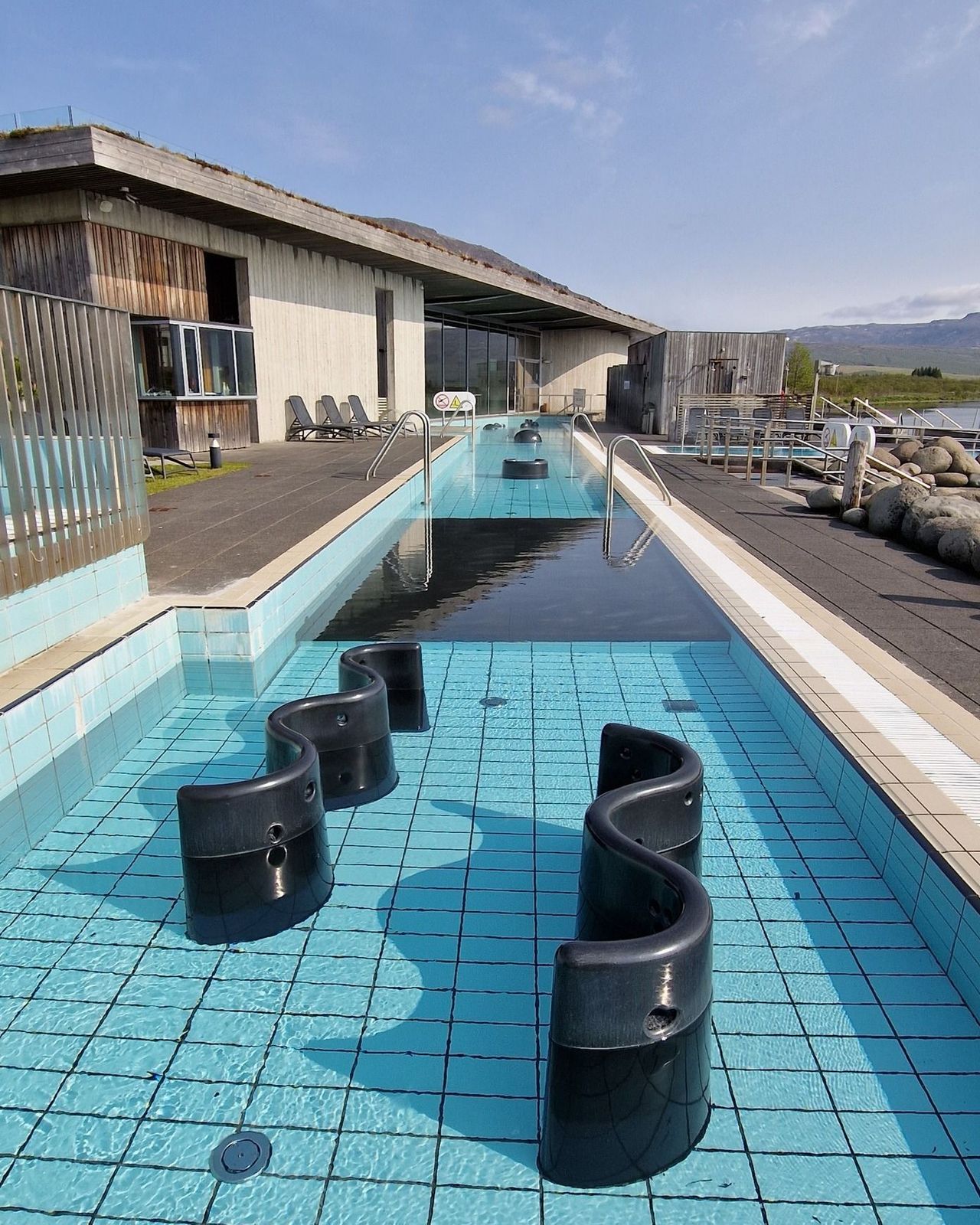 Outdoor swimming pool with S-shaped black barriers in the shallow end, a long lap pool, and a modern building with a green roof.