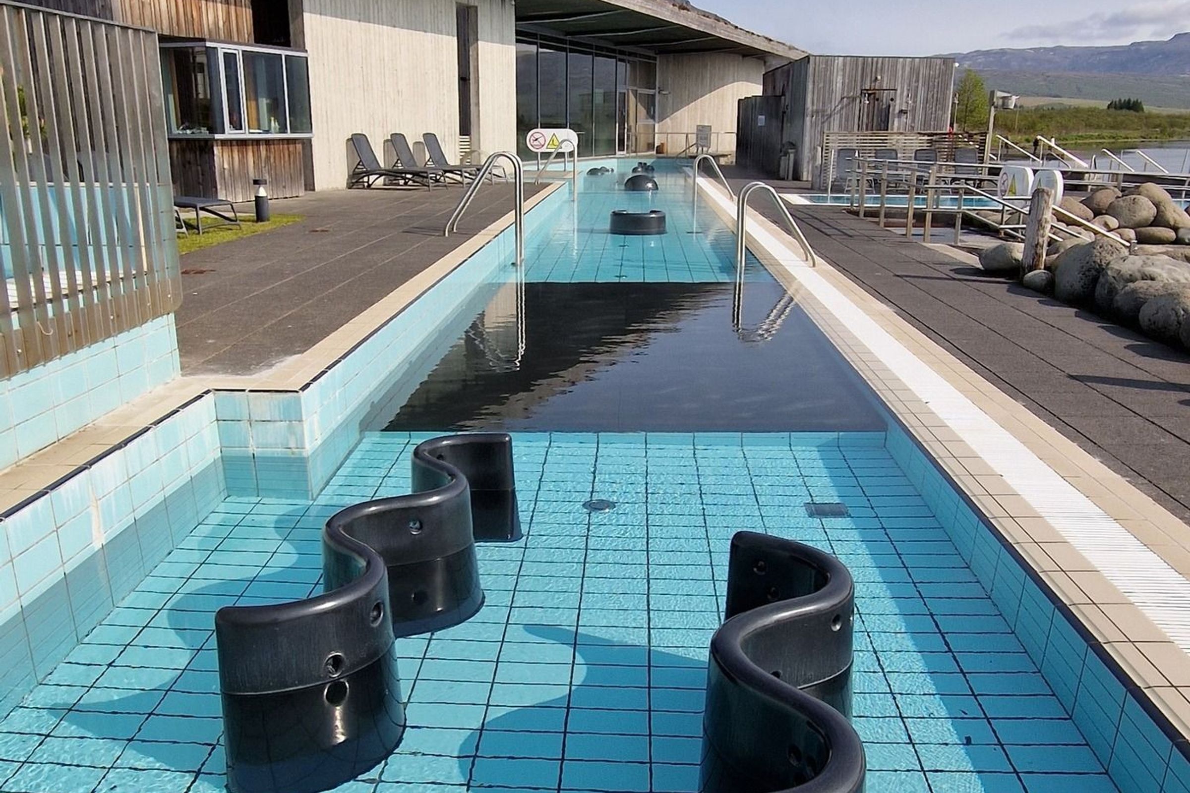 Outdoor swimming pool with S-shaped black barriers in the shallow end, a long lap pool, and a modern building with a green roof.