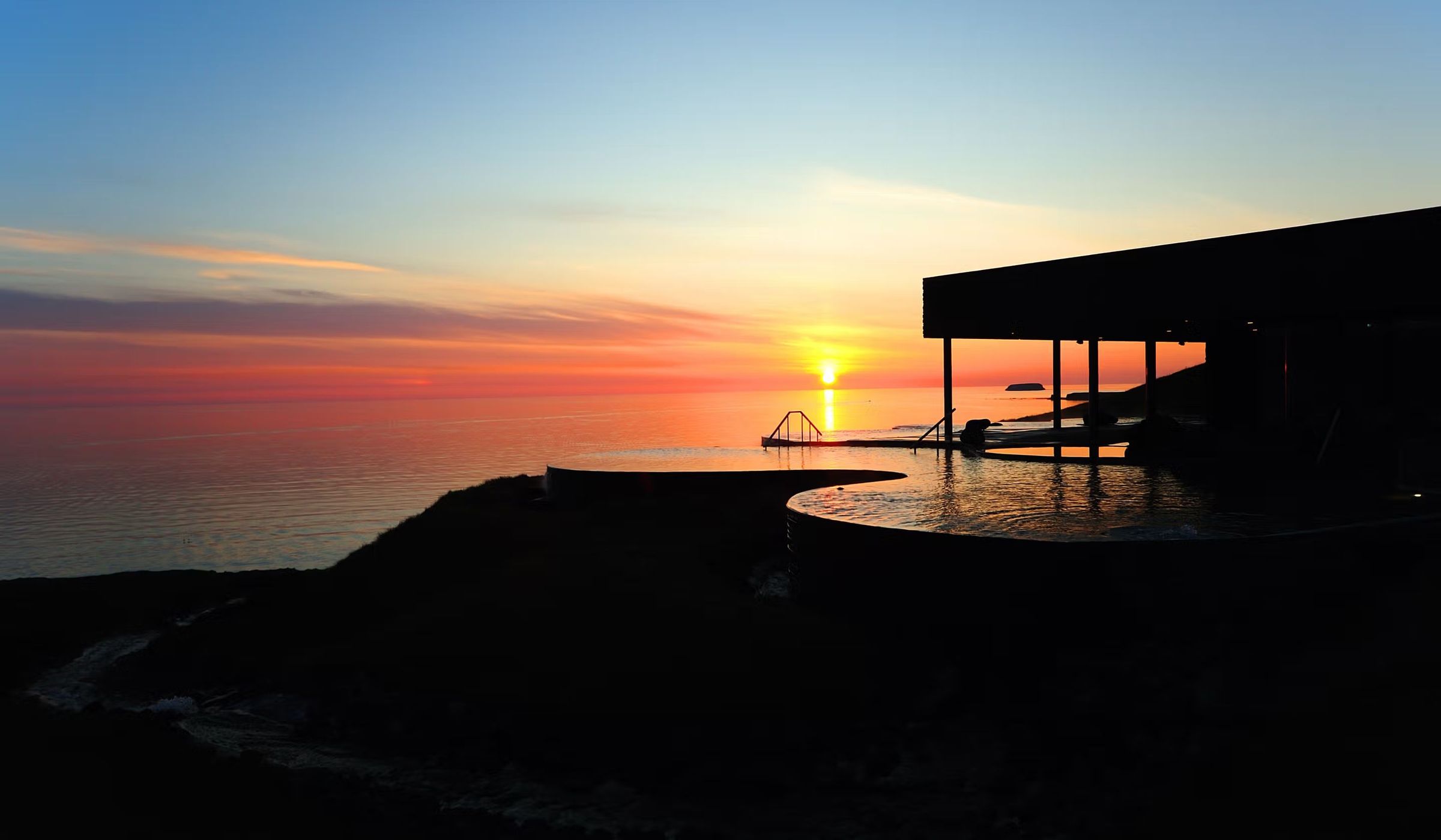 A modern building and outdoor pools silhouetted against a vibrant sunset over the ocean.