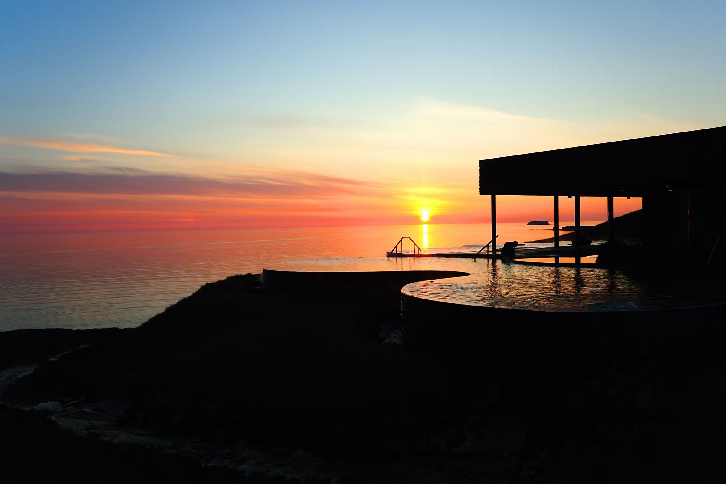 A modern building and outdoor pools silhouetted against a vibrant sunset over the ocean.