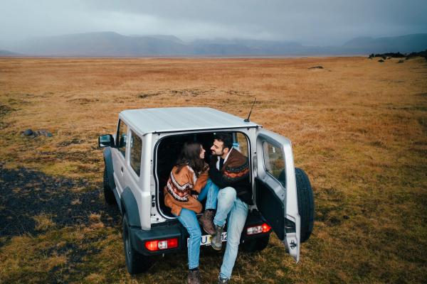 Una pareja está sentada en el maletero abierto de un SUV plateado, mirándose el uno al otro en medio de un campo vasto y seco de hierba, bajo un cielo nublado.