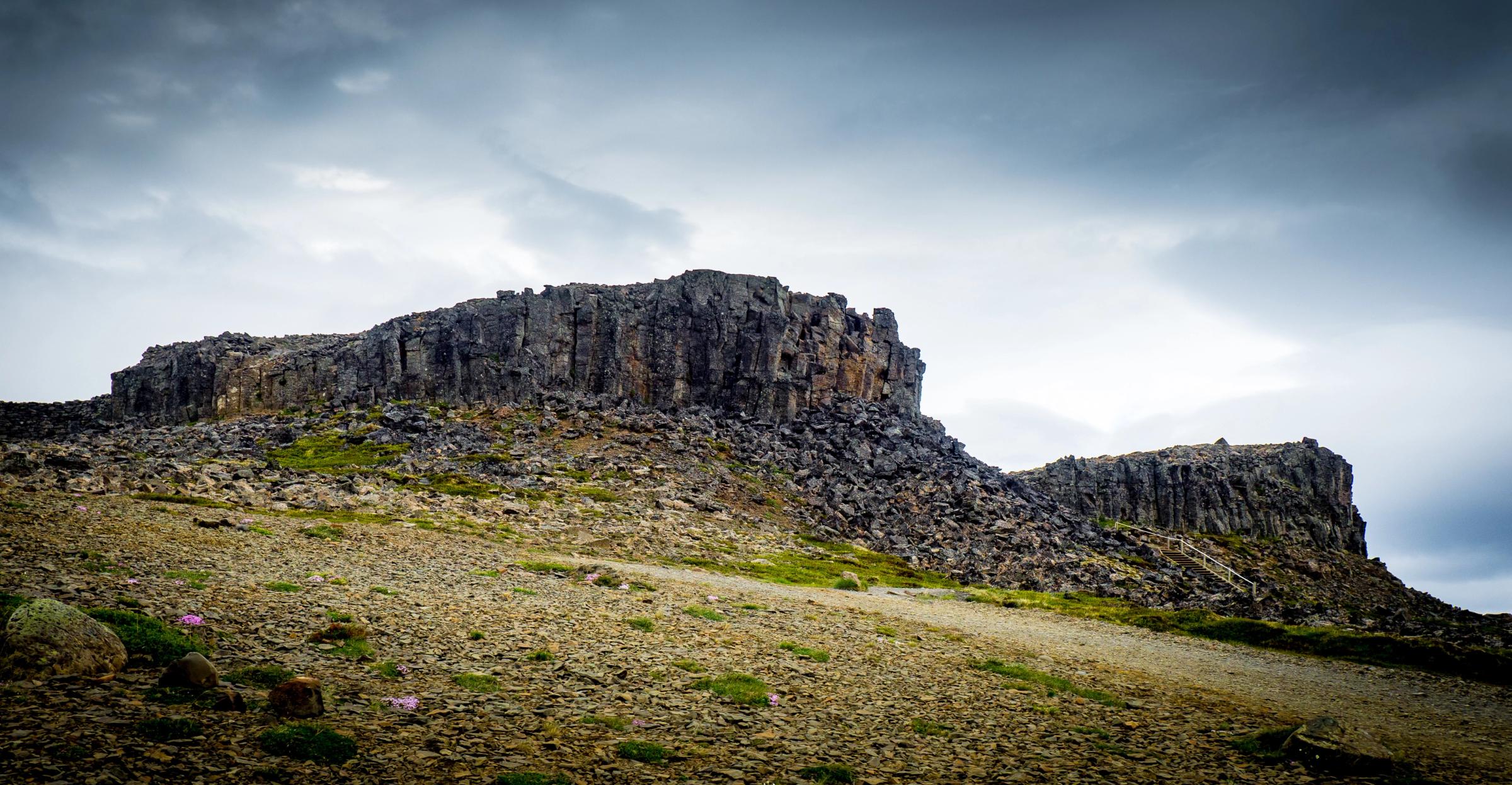 A rugged, rocky landscape with a dark cliff of columnar basalt formations under an overcast sky.