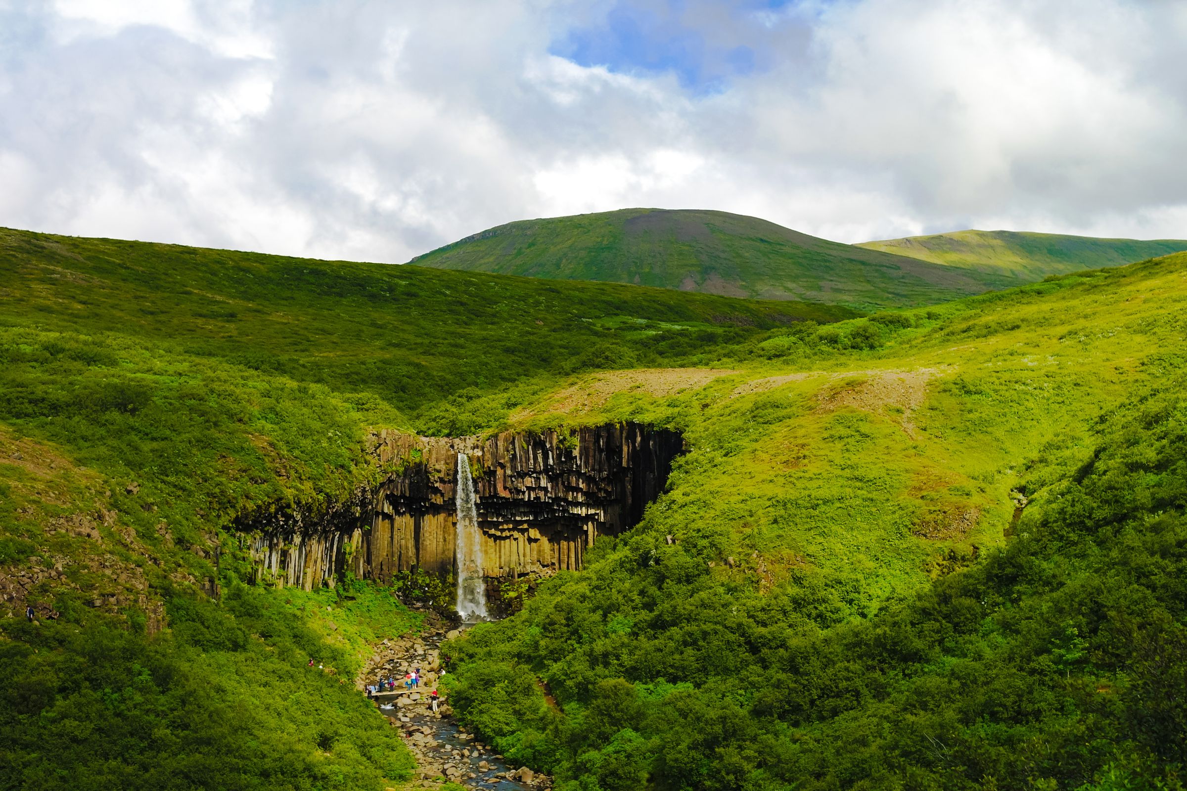 Una cascada se precipita sobre oscuras columnas de basalto, rodeada de colinas de un verde intenso bajo un cielo nublado.