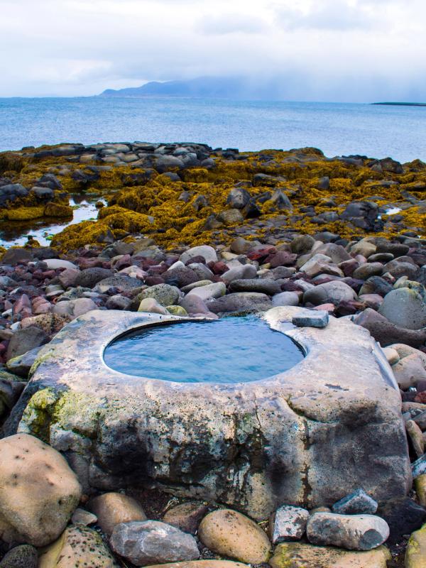 A circular hot spring pool built into a stone structure on a rocky beach, with the ocean and cloudy sky in the background.