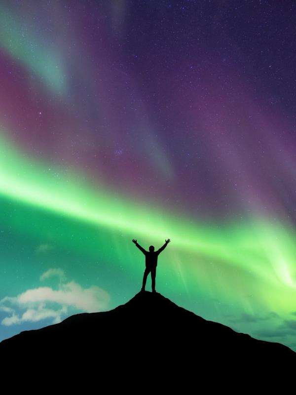 a man is standing on top of a hill with his arms outstretched in front of the aurora borealis .