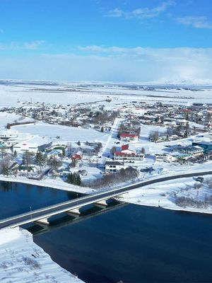 Aerial view of a snow-covered town next to a dark blue river with a bridge, under a blue sky, with mountains in the distance.