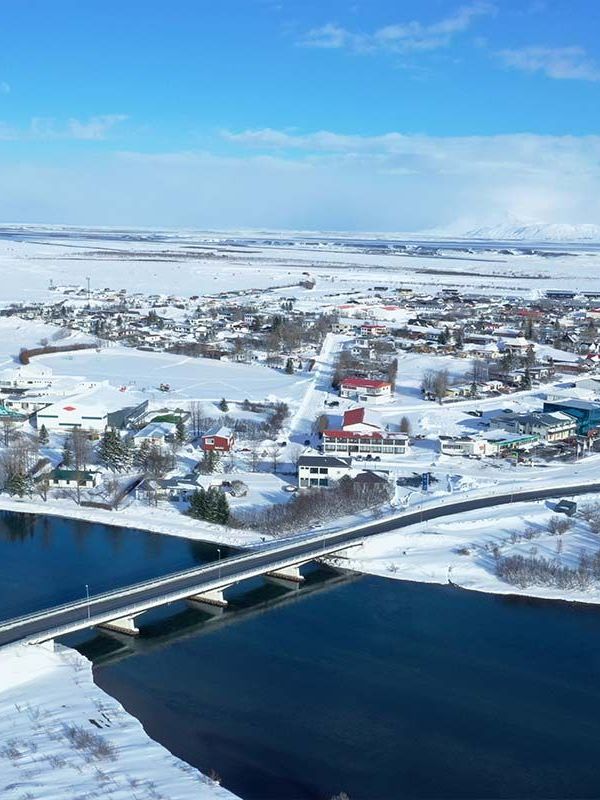 Vista aérea de un pueblo cubierto de nieve junto a un río de azul oscuro con un puente, bajo un cielo azul, con montañas en la distancia.