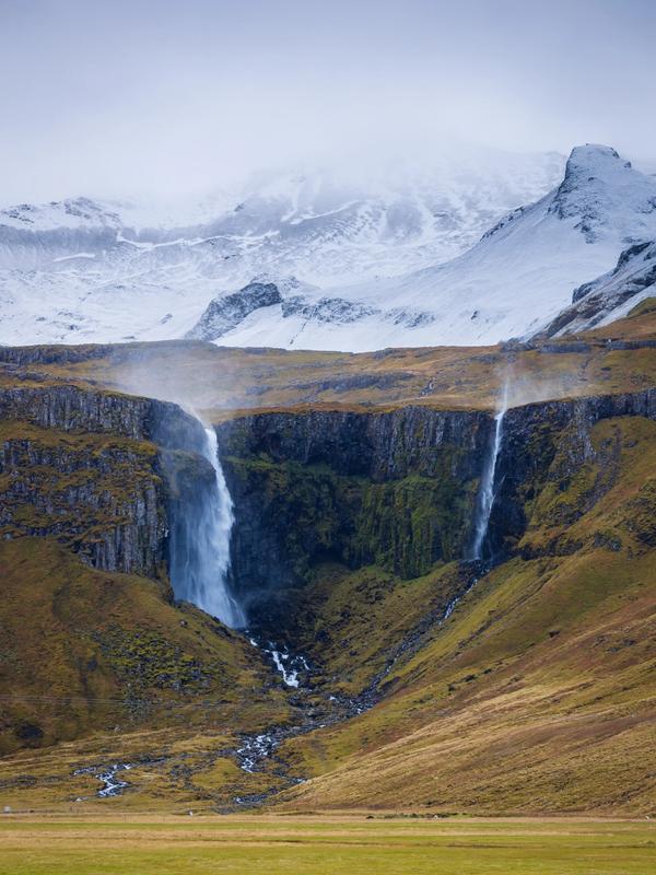 Two waterfalls cascade down a rocky cliff in a grassy valley, with snow-capped mountains in the background.