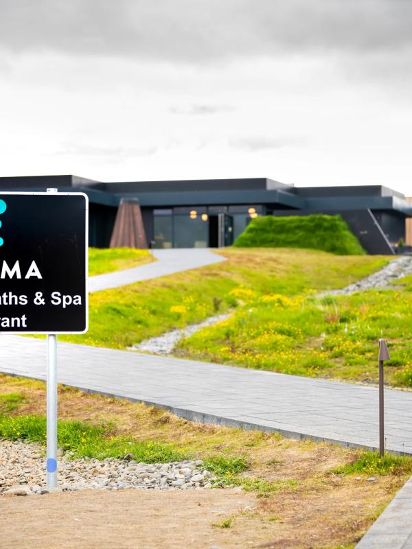 A sign for Krauma Geothermal Baths & Spa Restaurant stands by a pathway leading to a modern dark building with green landscaping.