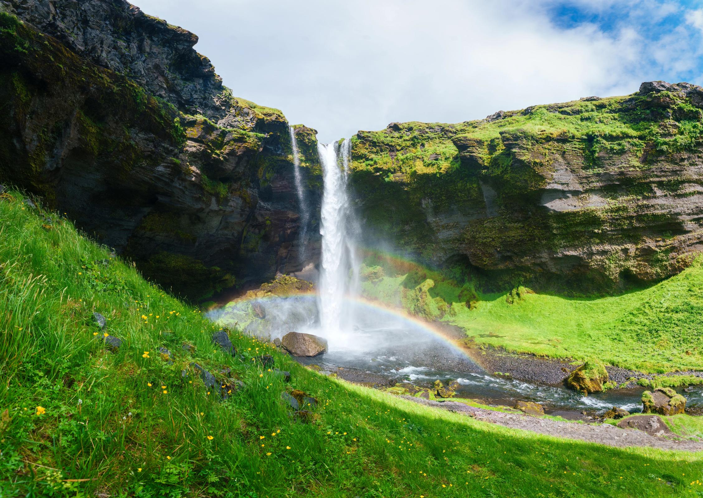 A waterfall descends over lush green cliffs with a rainbow arching through its mist.