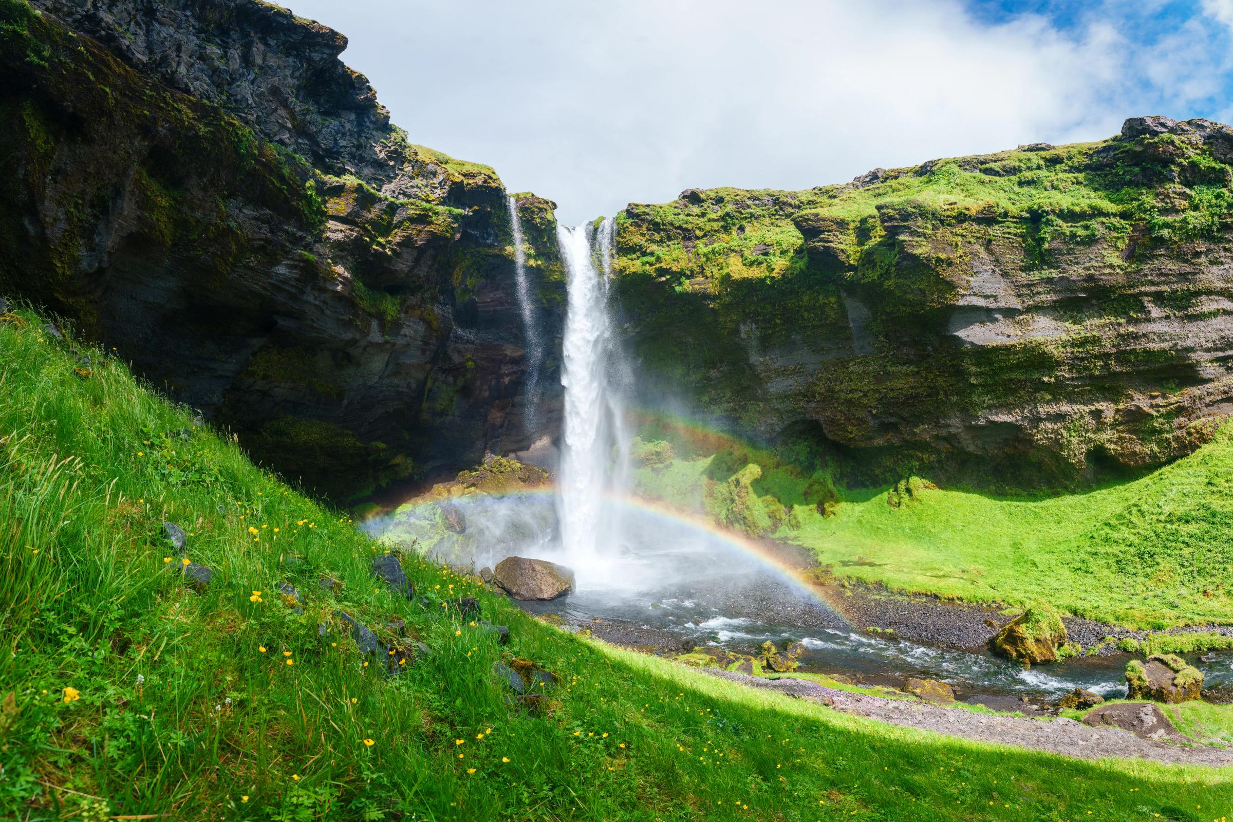 A waterfall descends over lush green cliffs with a rainbow arching through its mist.