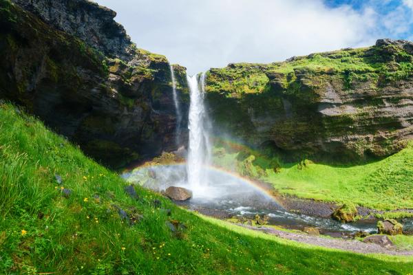 A waterfall descends over lush green cliffs with a rainbow arching through its mist.