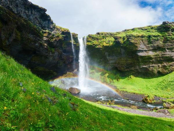A tall waterfall plunges over rocky green cliffs with a rainbow visible in the mist below, surrounded by lush grass.