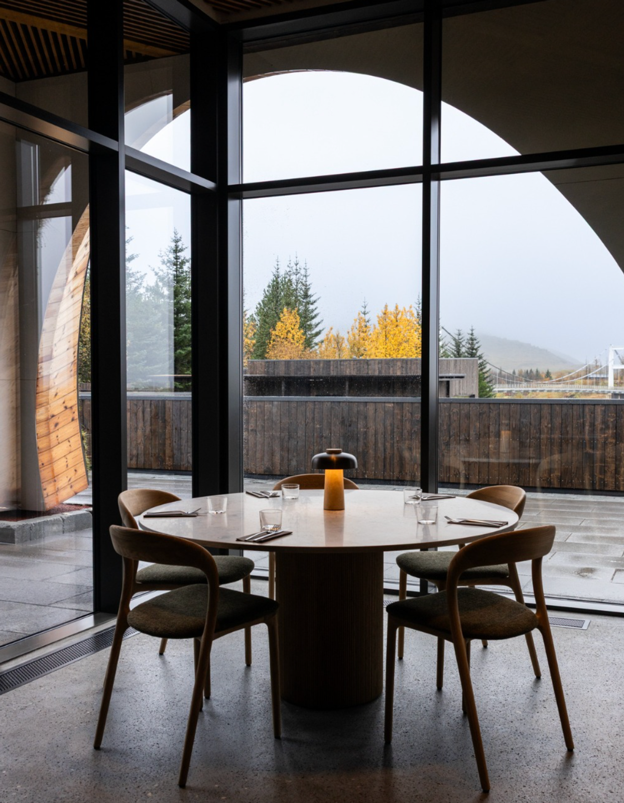 A modern restaurant interior with a round table set for four and a glowing lamp, looking out at autumn trees.