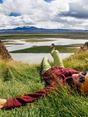 Chica con ropa alegre, gorro, y gafas en el suelo con un lago al fondo