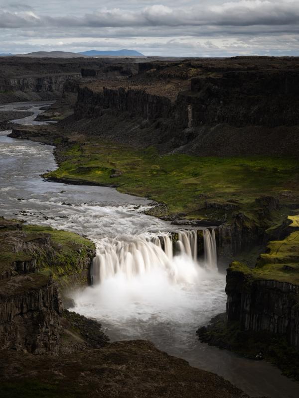 A wide waterfall plunges into a deep, dark canyon with green patches, under a cloudy sky.