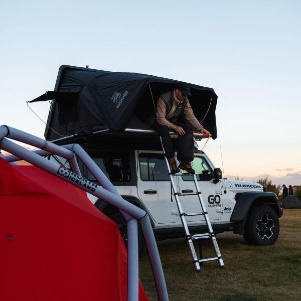 Un chico sentado encima de un jeep blanco que tiene una tienda de campaña en el techo