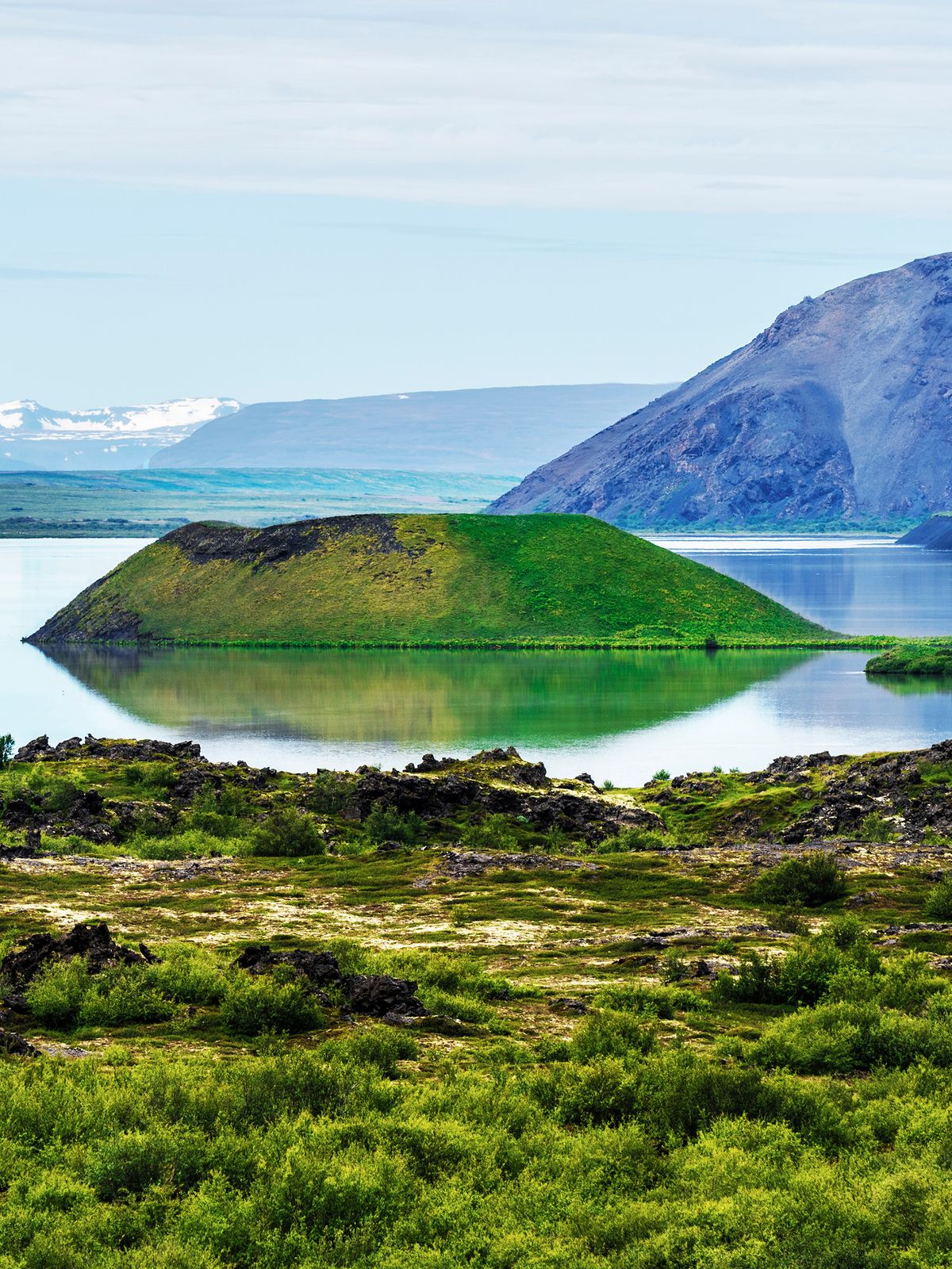 Volcanic landscape with a lake, green pseudo-craters, a large dark mountain, and distant snowy peaks.