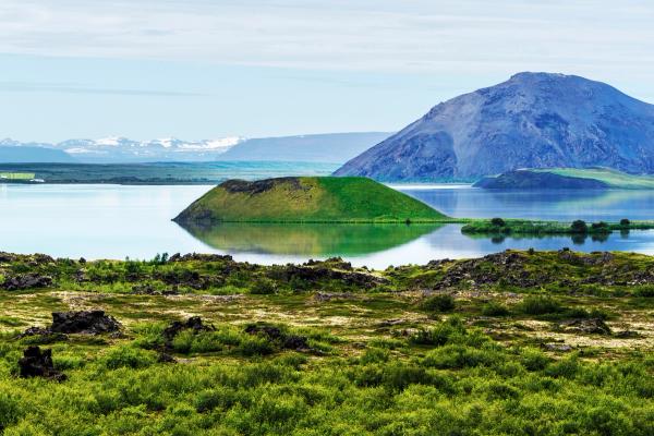 A calm lake reflecting a vibrant green conical hill, with a large dark mountain and distant snow-capped peaks in the background, and rocky green terrain in the foreground.