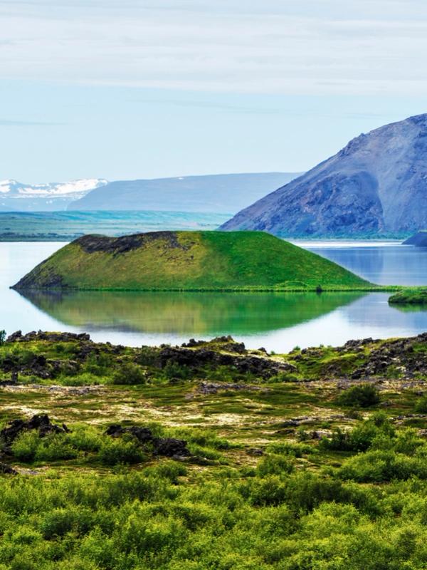 Volcanic landscape with a lake, green pseudo-craters, a large dark mountain, and distant snowy peaks.