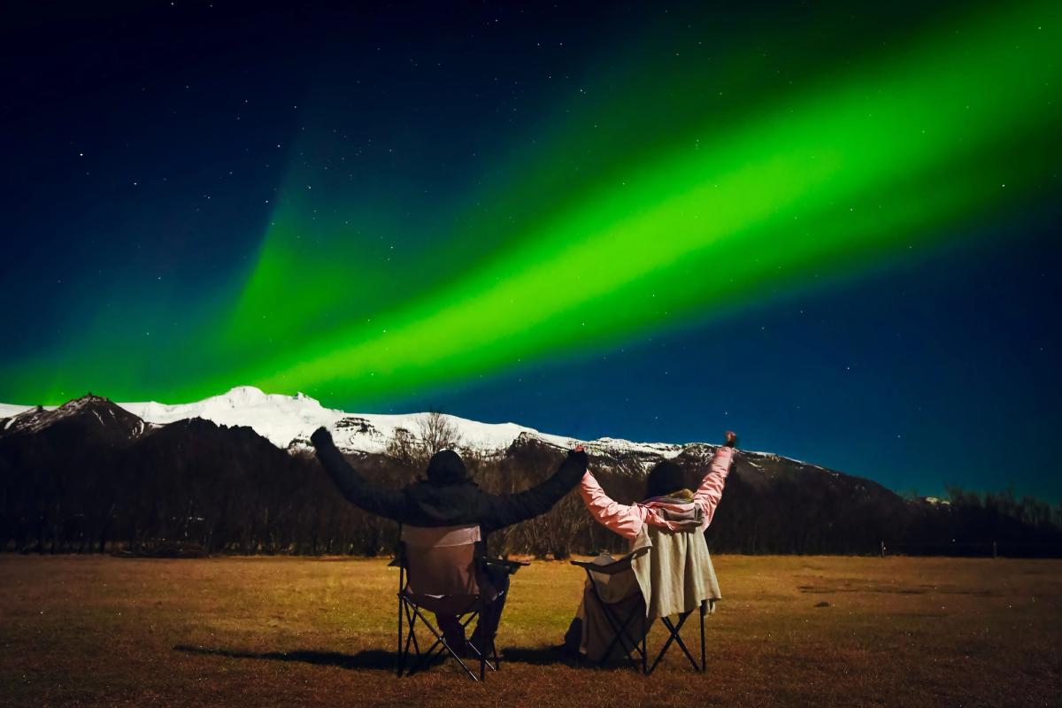 Two people with raised arms watch vibrant green Northern Lights over snow-capped mountains.