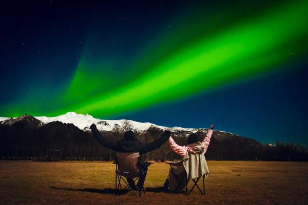 Two people with raised arms watch vibrant green Northern Lights over snow-capped mountains.