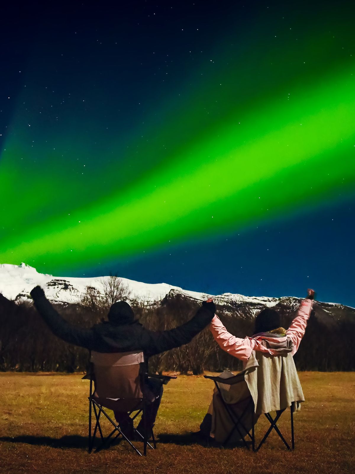 Two people with raised arms watch vibrant green Northern Lights over snow-capped mountains.