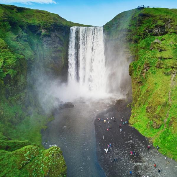 A powerful waterfall cascades from green cliffs, with many people on the dark shore at its base.